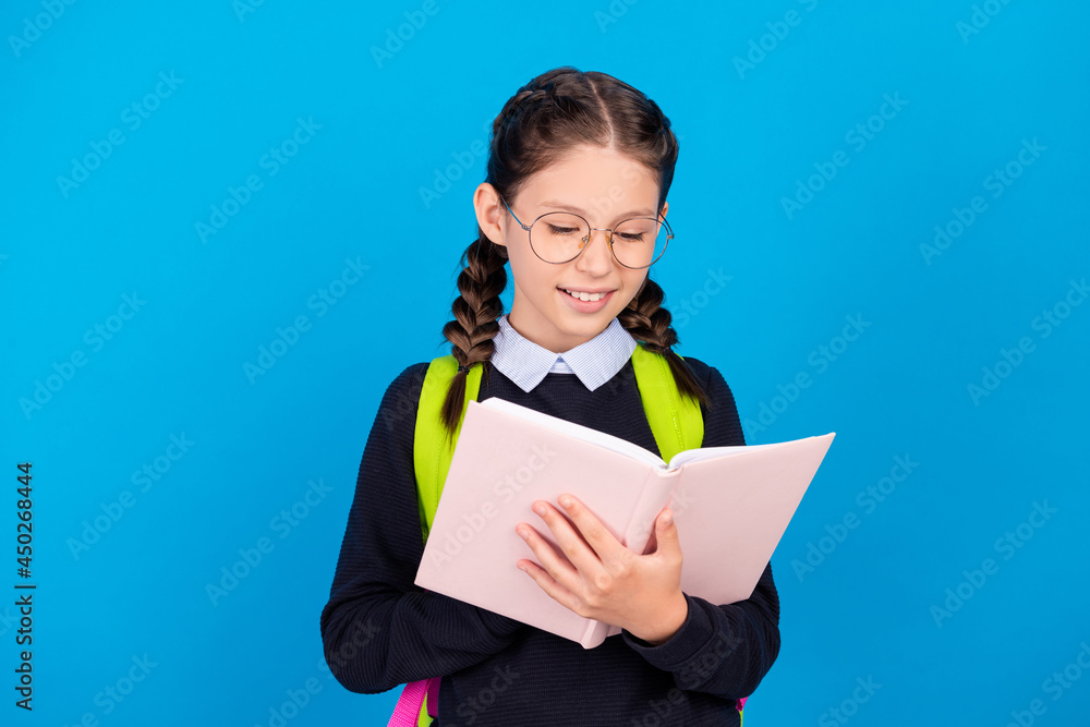 Photo of happy smiling young schoolgirl in spectacles reading book prepare homework isolated on blue color background Photo of happy smiling young schoolgirl in spectacles reading book prepare homework isolated on blue color background