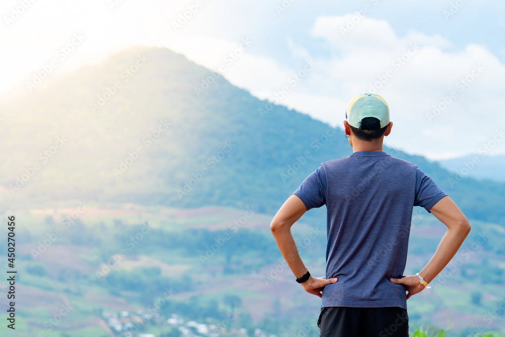 Half-body shot of an Asian male runner in a blue shirt and hat standing warm up in preparation for a jogging run. It is located on a high cliff with beautiful mountains in the background.