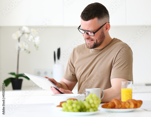 Young smiling handsome man working remotely on tablet in modern kitchen at home