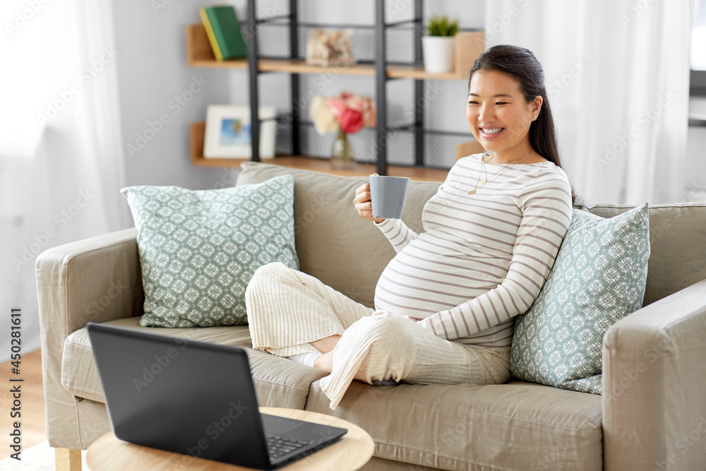 pregnancy, rest, people and expectation concept - happy smiling pregnant asian woman with laptop computer sitting on sofa at home and drinking tea pregnancy, rest, people and expectation concept - happy smiling pregnant asian woman with laptop computer sitting on sofa at home and drinking tea