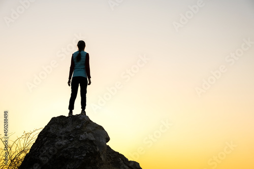 Silhouette of a woman hiker standing alone on big stone at sunset in mountains. Female tourist on high rock in evening nature. Tourism, traveling and healthy lifestyle concept.