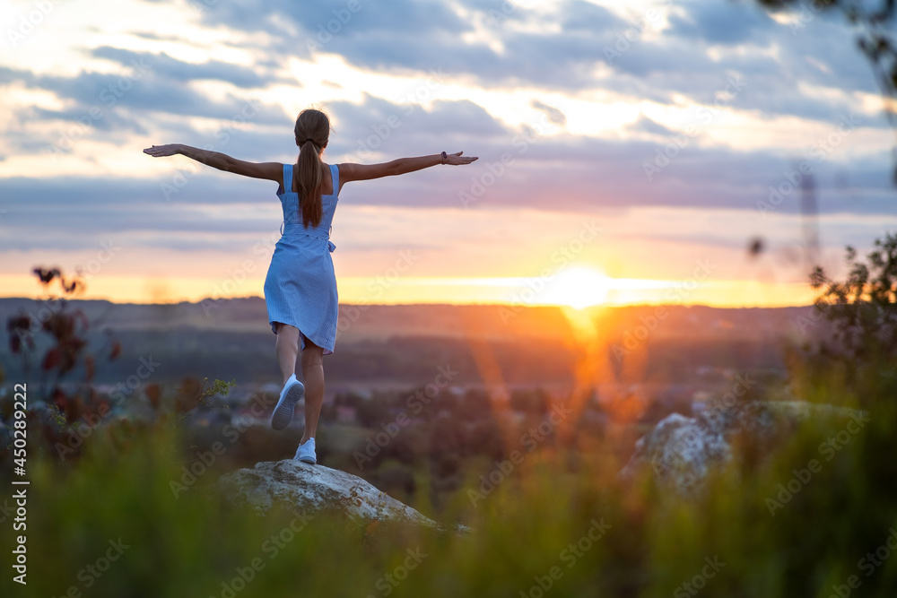 A young woman in summer dress standing outdoors with outstretched arms enjoying view of bright yellow sunset.