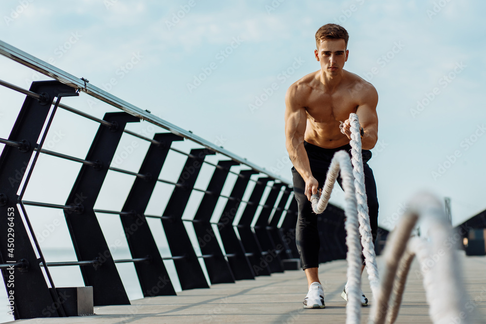 Naklejka premium Strong man training with fighting ropes, Sportsman doing battle rope training outdoors on the sea on the pier