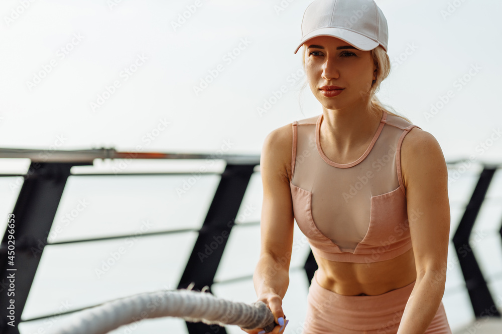 Fototapeta premium Fitness woman exercising with fighting ropes outdoors on the pier, woman in sportswear doing fight ropes workout