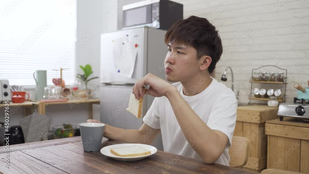 carefree young Asian man is biting into the toast and drinking coffee from cup while enjoying the first meal of the day on a tranquil morning at home. carefree young Asian man is biting into the toast and drinking coffee from cup while enjoying the first meal of the day on a tranquil morning at home.
