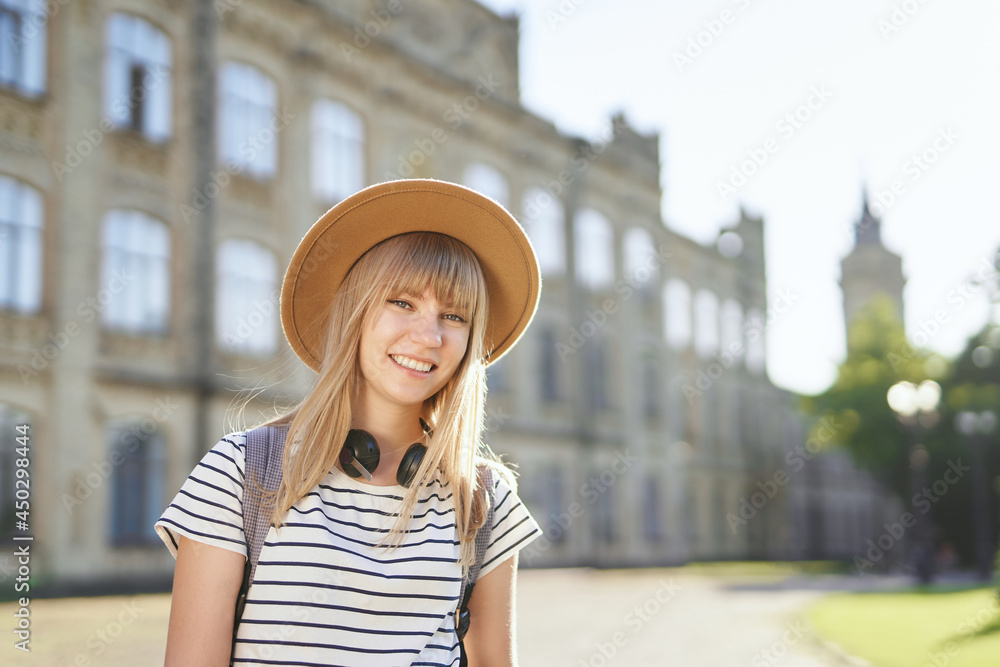Cheerful smiling young blonde female European university or college student wearing brown hat in campus. Cute school girl, student portrait or education concept. High quality image