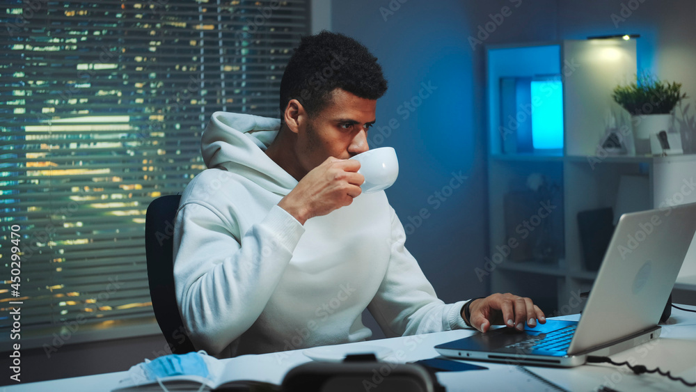Handsome multiracial man drinking a cup of coffee and working on computer at night. There are skyscrapers in the background.