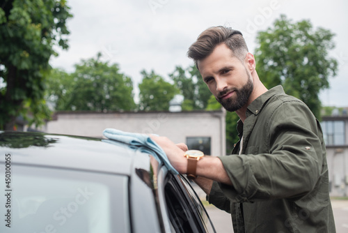 Bearded driver cleaning blurred car with rag outdoors