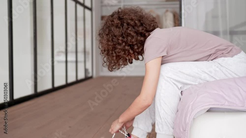 young woman with curly hair sitting on bed preparing for morning jogging ties shoelaces bright running sneakers everyday sport routine