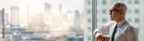 thoughtful senior business man holding cup of coffee standing over window glass in office on high building cityscape view background . worry old worker distracted . executive thinking and consider