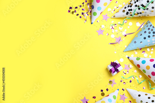 Flat lay composition with party hats, gift box and confetti on yellow background, space for text. Birthday decor