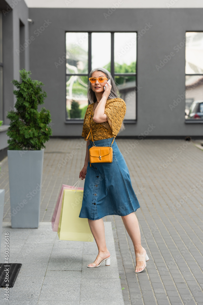stylish middle aged woman with shopping bags talking on mobile phone near building