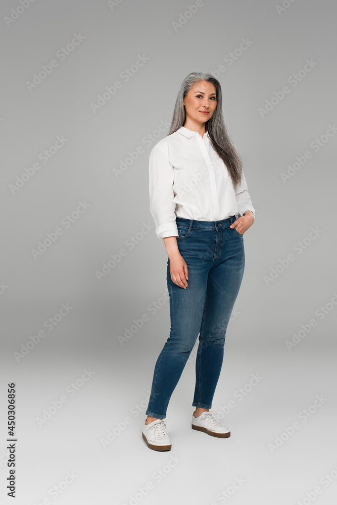 full length view of smiling middle aged woman in blue jeans and white shirt on grey full length view of smiling middle aged woman in blue jeans and white shirt on grey