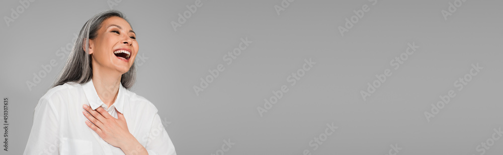 cheerful middle aged woman in white shirt laughing with closed eyes isolated on grey, banner