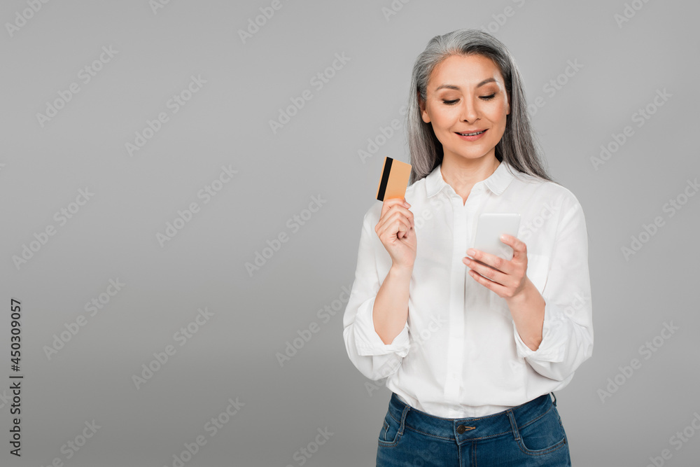 Fototapeta premium smiling woman in white shirt holding credit card while looking at mobile phone isolated on grey