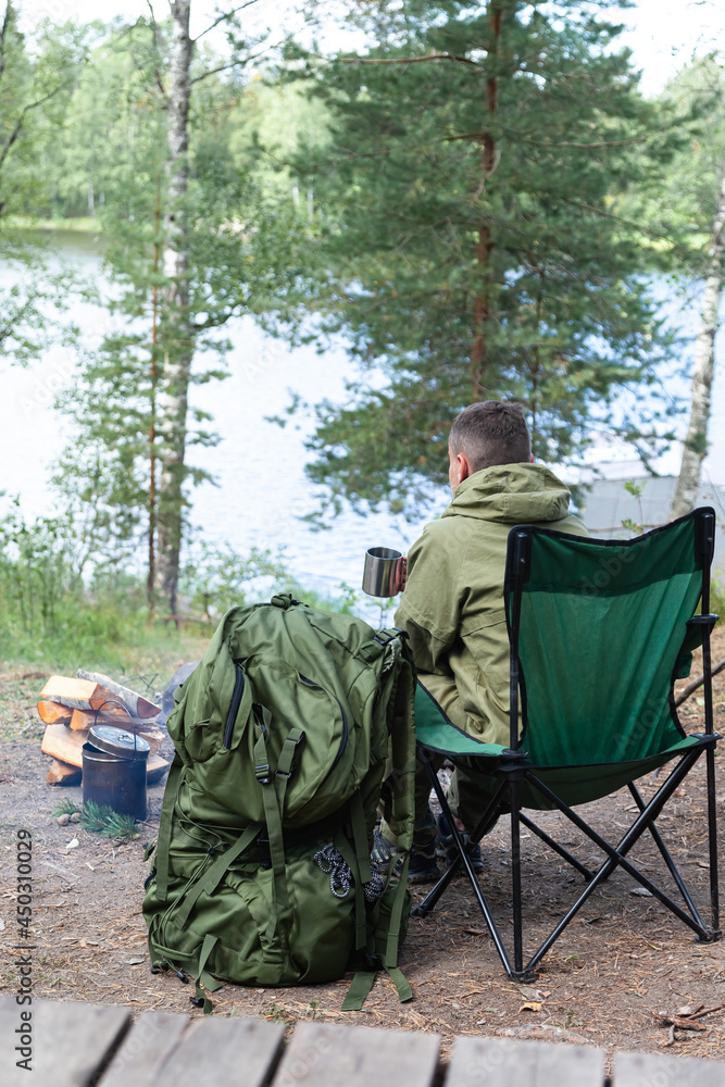 Young man travelling alone in national parks, drinking warm tea near the bonfire from metal mug. Concept of active tourism, camping in the forecast, trekking and hiking during vacations. Close up