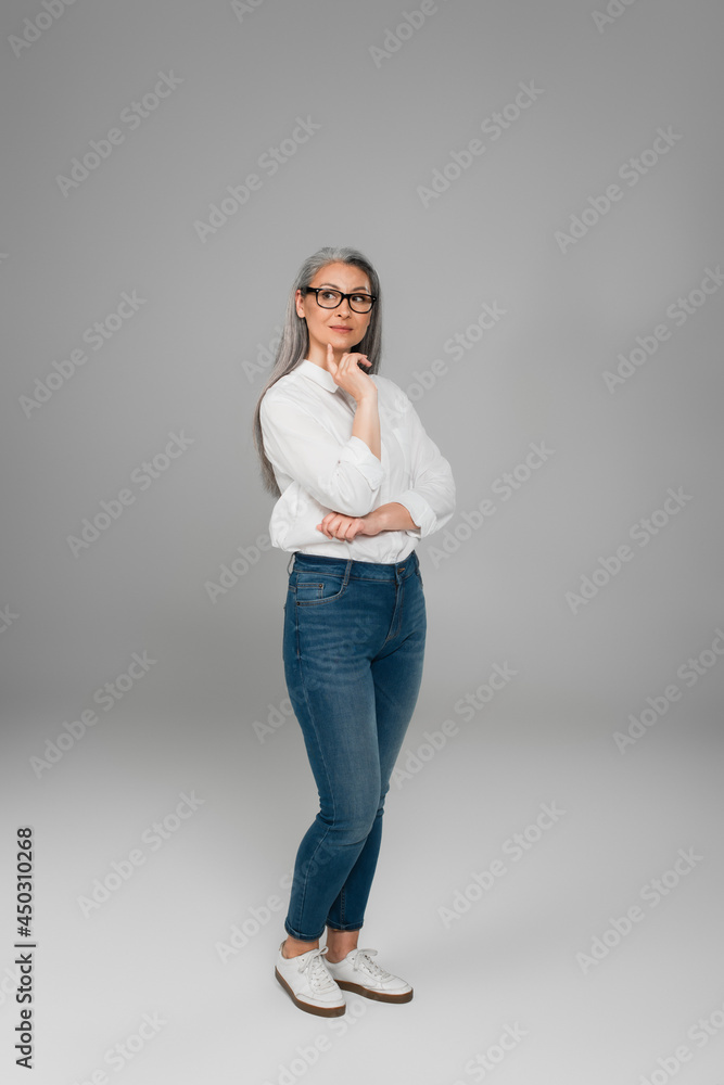 full length view of mature woman in jeans, white shirt and eyeglasses standing on grey full length view of mature woman in jeans, white shirt and eyeglasses standing on grey
