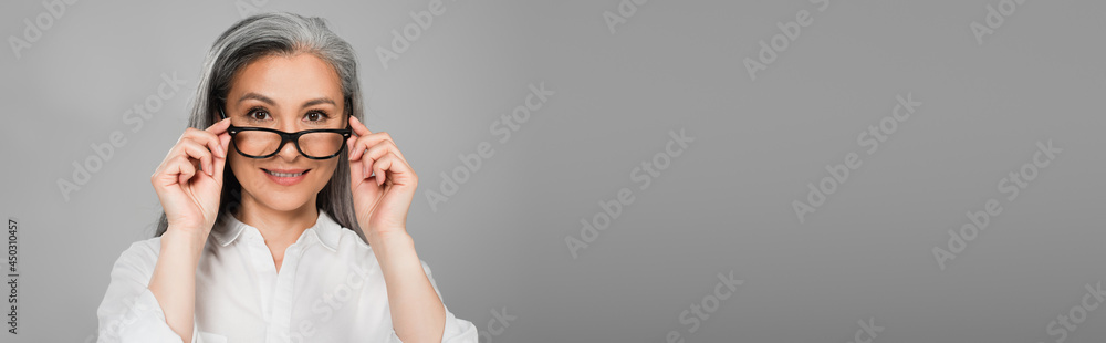 positive, grey haired woman smiling at camera while taking off eyeglasses isolated on grey, banner