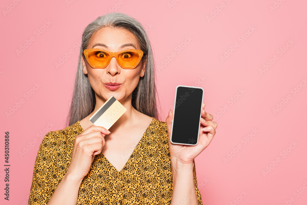 excited mature woman with credit card showing mobile phone with blank screen isolated on pink