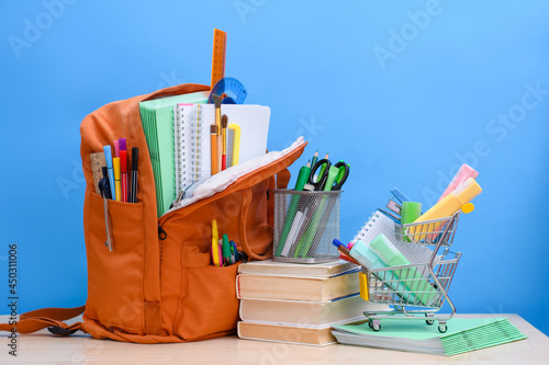 Orange school backpack full of school supplies and a supermarket basket with office supplies on a blue background. The concept of gathering children for the beginning of the school year.