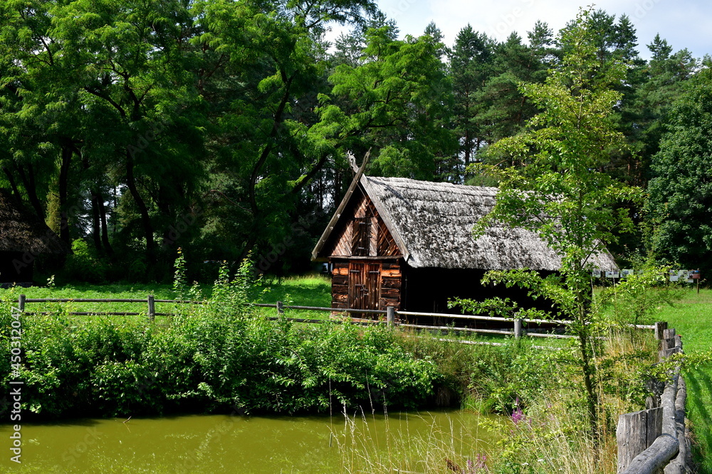 A close up on a wooden shack, hut, or house standing next to a small pond, lake, or swamp covered with shrubs, reeds and other flora seen on a sunny summer day near a dense forest or moor in Poland A close up on a wooden shack, hut, or house standing next to a small pond, lake, or swamp covered with shrubs, reeds and other flora seen on a sunny summer day near a dense forest or moor in Poland