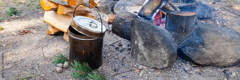 Metal bowler pot near bonfire and stake of wood. Preparation of soup or tea. Concept of traveling, active tourism, picnic cooking on fire. Close up banner