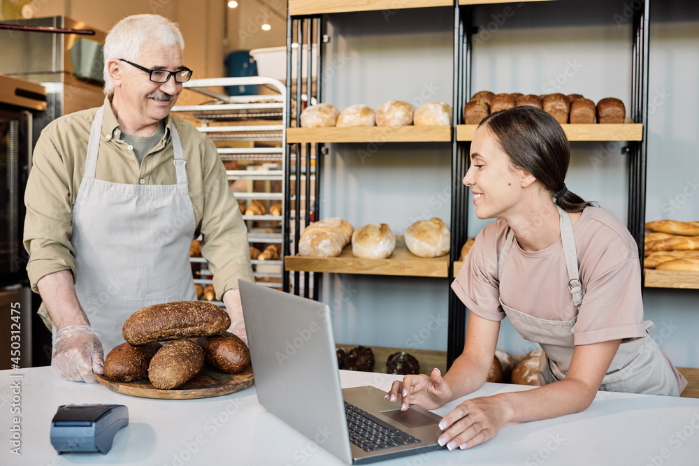 Happy woman with laptop looking at pile of fresh loaves of bread on wooden board held by mature baker Happy woman with laptop looking at pile of fresh loaves of bread on wooden board held by mature baker