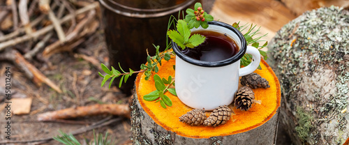 White campfire enamel mug with hot herbal tea on wooden stump. Bowler pot, backpack, cones, forest on background. Concept of lunch break during hiking. Active tourism, camping. Banner copy space
