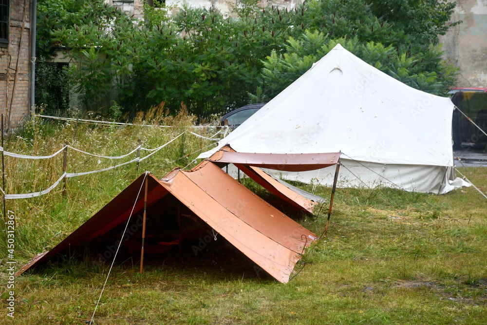 A close up on two military tents made out of sturdy cloth and supported with planks, sticks, and ropes standing in the middle of a field overgrown with grass and shrubs seen right after a heavy rain