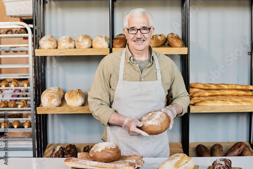 Happy aged baker with loaf of fresh bread looking at you by workplace