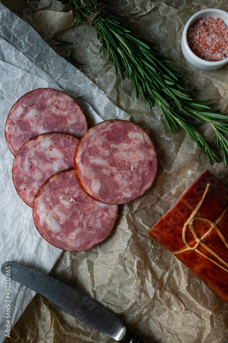 Slices of round sausage on parchment paper with silver knife, pink salt and rosemary twigs