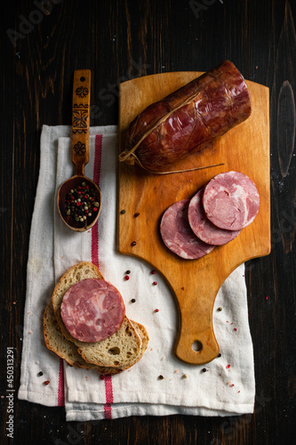 Slices of salami sausage on a wooden cutting board  with assorted peppercorns in a wooden spoon and  a slice of white bread on white kitchen towel and dark wooden table