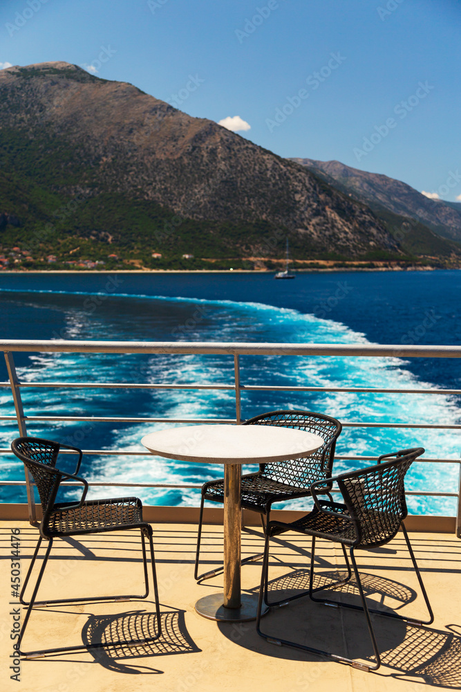 Table with three chairs at cruise ship open deck with mountain landscape of Kefalonia island. Ferry boat leaving Cephalonia island.
