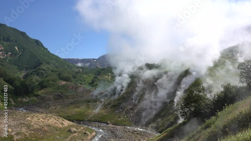 Valley of Geysers. Kamchatka. Steam rises from several hot springs over the mountainside. Fog against the blue sky. In the valley, a river flows along a rocky bed. A sunny summer day