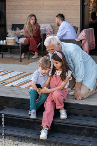 Two kids and their grandfather talking through video chat on staircase against young couple © pressmaster