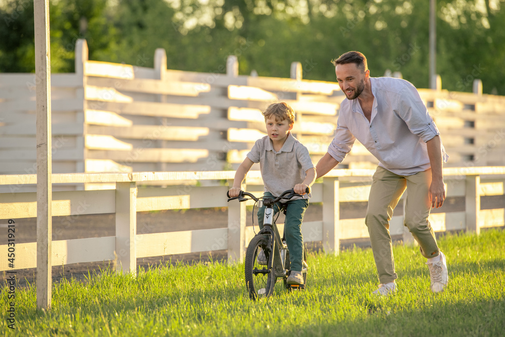 Happy young man helping his little son to ride bicycle along green lawn on sunny day Happy young man helping his little son to ride bicycle along green lawn on sunny day