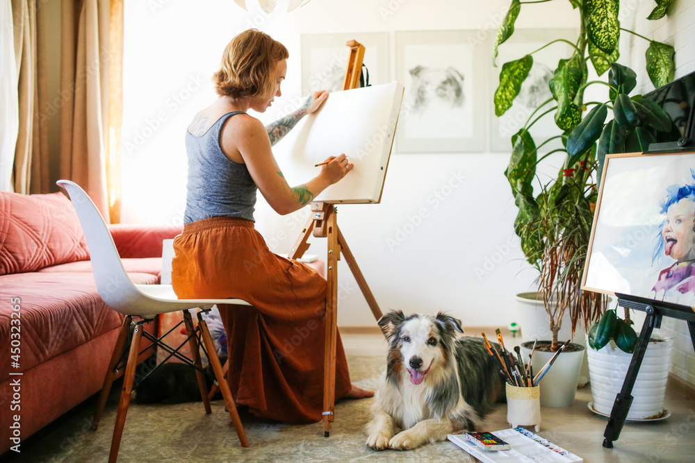 A cute red-haired girl artist draws together with the Aussie dog, Australian Shepherd breed. Owner and pet together in a bright living room, home workshop