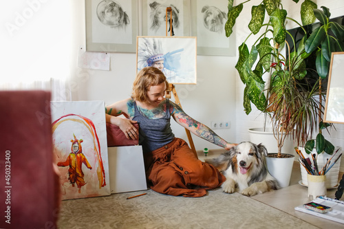 A cute red-haired girl artist paints together with the Aussie dog, the Australian Shepherd breed. Owner and pet together in a bright living room, home workshop, lifestyle in real room