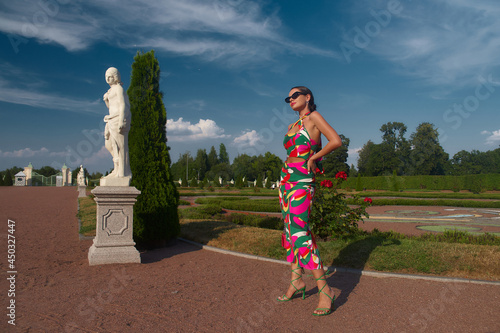 Stylish elegant woman in colorful vivid pink green dress, heels and sunglasses standing and posing in beautiful garden with flowers and sculptures. Fashion full length portrait photo