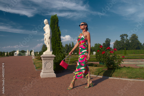 Stylish elegant woman in colorful vivid dress, heels and sunglasses with pink handbag walking in beautiful garden with flowers and sculptures. Fashion full length portrait photo