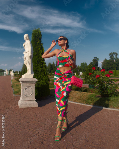 Stylish elegant woman in colorful vivid dress, heels and sunglasses with pink handbag standing and posing in beautiful garden with flowers and sculptures. Fashion full length portrait photo