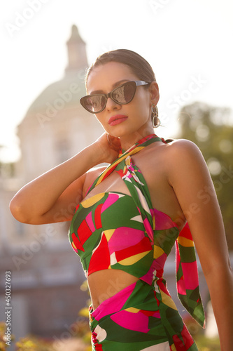 Close-up fashion portrait of elegant tanned caucasian woman in colorful stylish dress and sunglasses posing in palace garden on a sunny evening photo