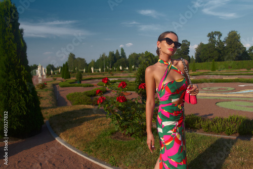 Stylish elegant woman in colorful vivid dress, heels and sunglasses with pink handbag standing and posing in beautiful garden with flowers. Fashion full length portrait photo