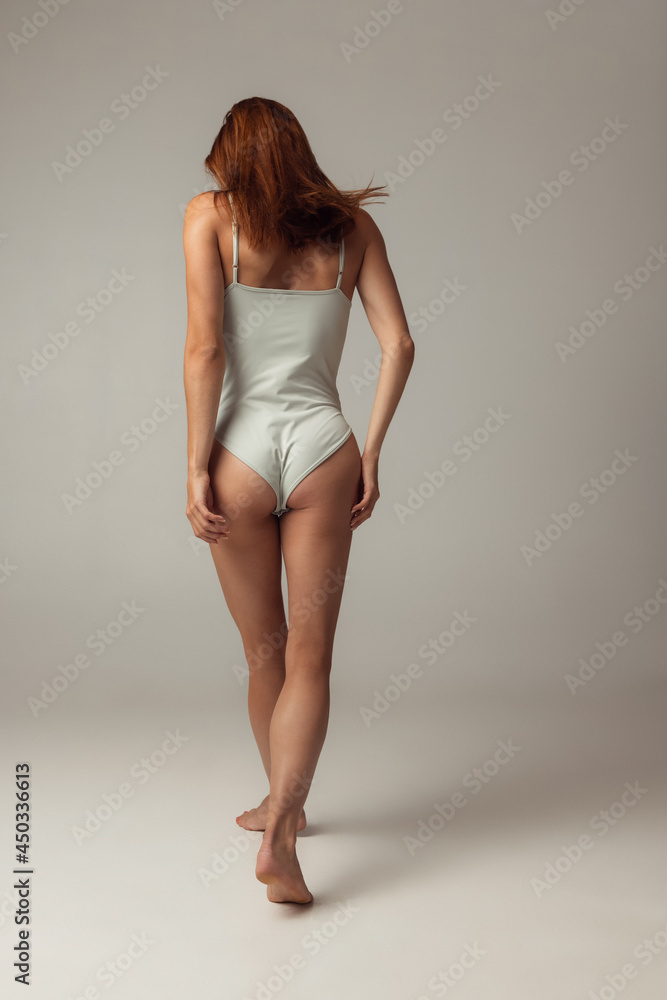 Portrait of young beautiful tanned red headed woman in lingerie posing isolated over gray studio background. Natural beauty concept. Back view