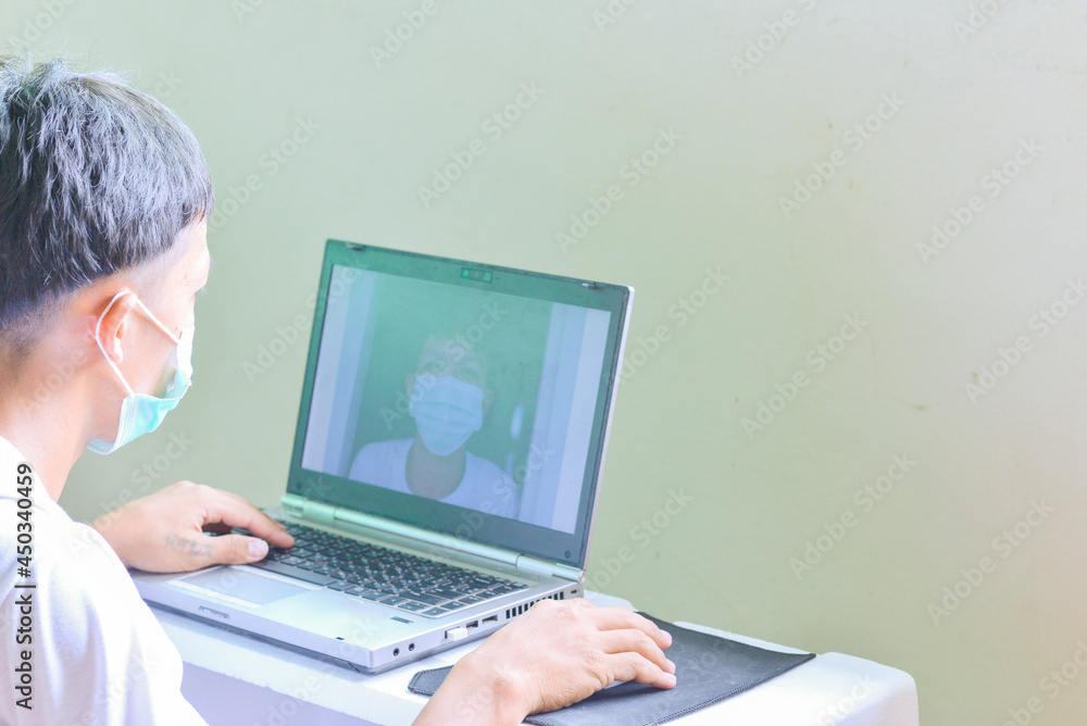 Asian man wearing a mask and video calling his friend on a laptop Asian man wearing a mask and video calling his friend on a laptop