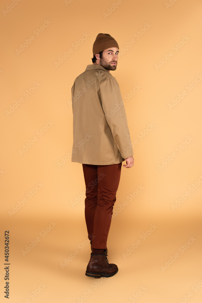 full length view of fashionable man in beanie, jacket and brown pants looking at camera on beige