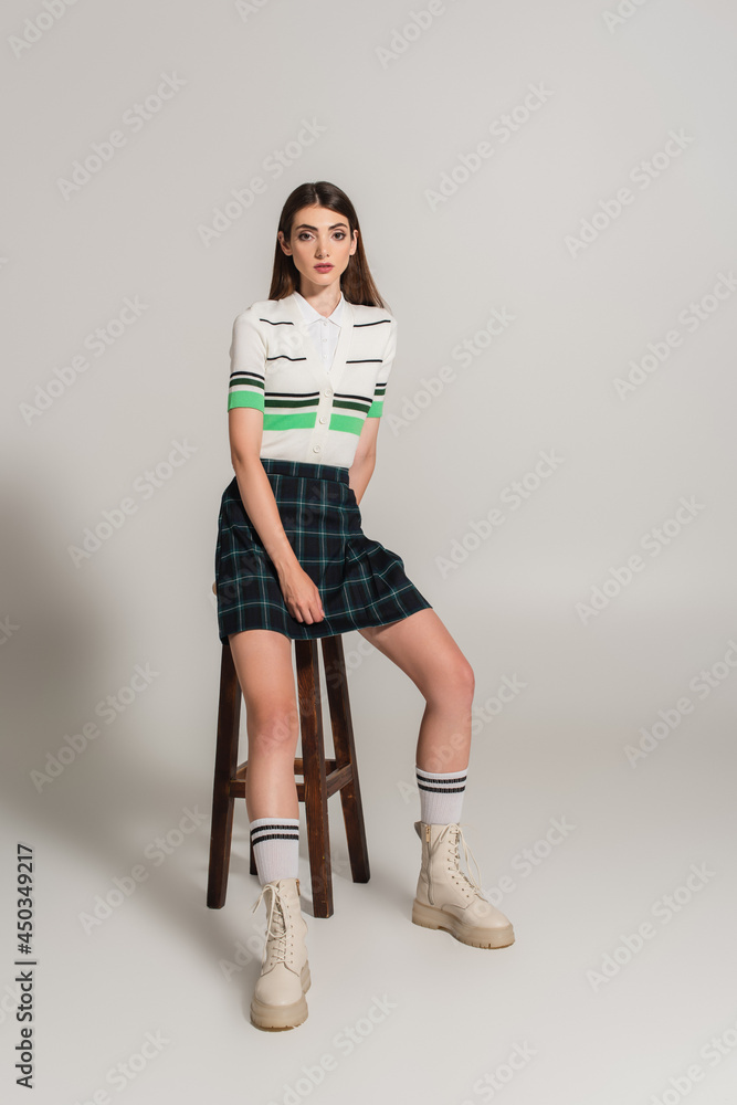 young woman in trendy clothes sitting on high wooden stool on grey background