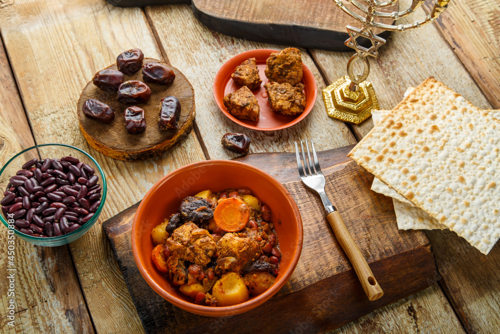 Jewish dish chelnt with meat on a stand next to matzo, fork and ingredients.