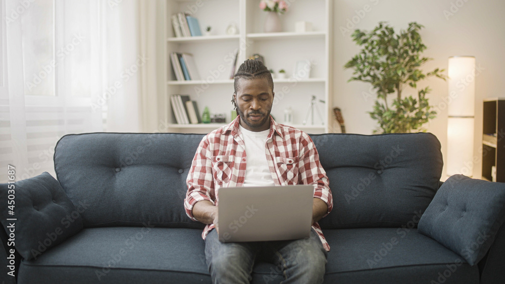 Young Afro-american man sitting on couch with laptop, working remotely, using apps Young Afro-american man sitting on couch with laptop, working remotely, using apps