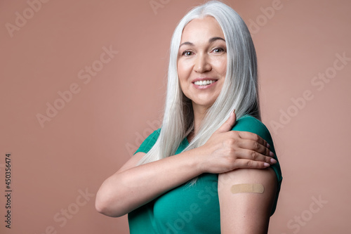 Happy vaccinated mature female showing shoulder with adhesive plaster after immunization vaccine. Smiling senior woman after receiving corona virus vaccination, studio portrait with copy space. photo